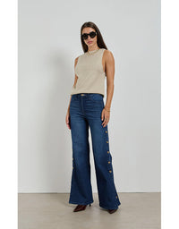 Woman wearing a beige sleeveless top and blue jeans with button details against a white background