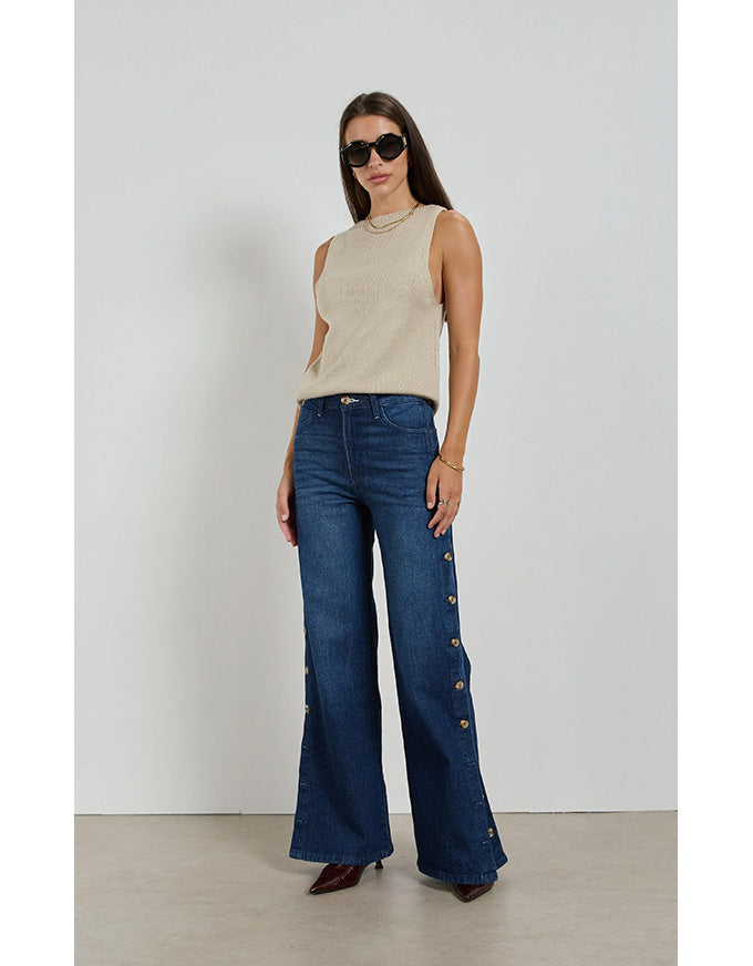 Woman wearing a beige sleeveless top and blue jeans with button details against a white background