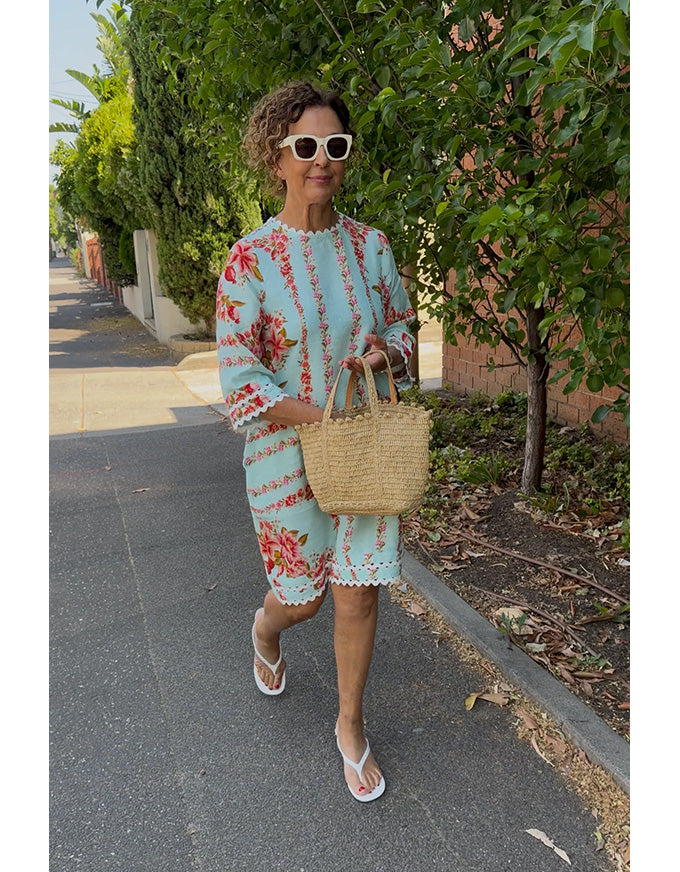 Woman in a floral dress holding a straw bag on a sidewalk with greenery.