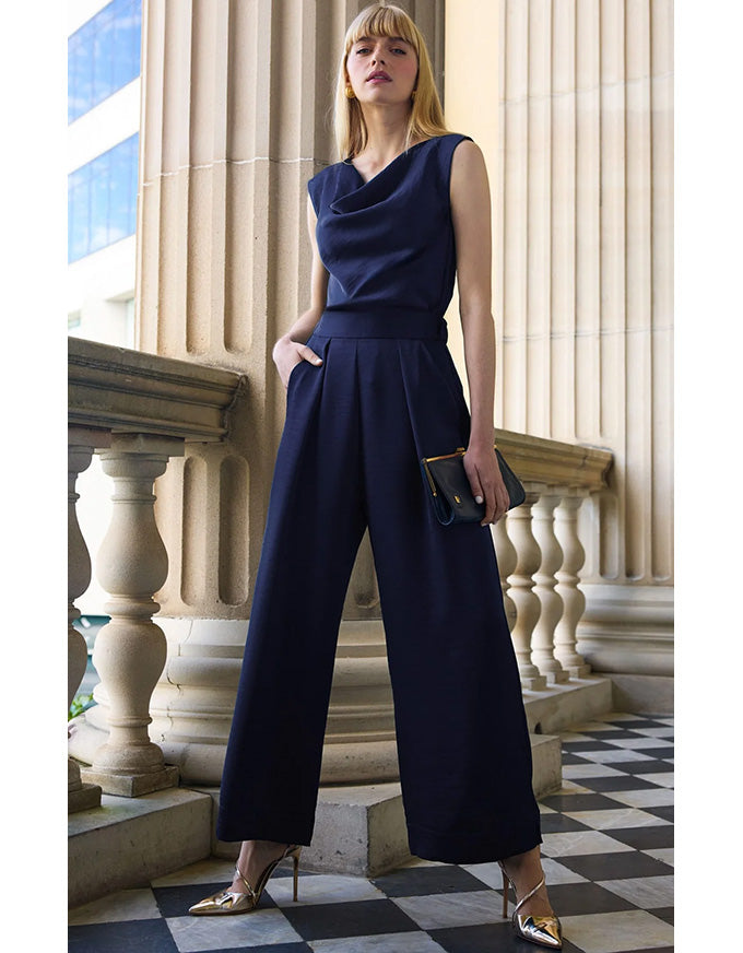 A woman models a navy blue sleeveless wide-leg jumpsuit in an elegant outdoor setting with stone columns and a checkered floor.