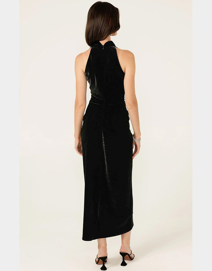 A woman viewed from the back wearing a sleeveless black halter-neck midi dress with a matching belt, photographed against a plain light beige studio background.