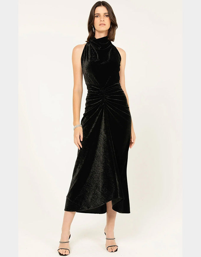 A woman modeling a sleeveless black velvet midi dress with a high mock neck, gathered waist, and draped skirt, set against a plain light background.