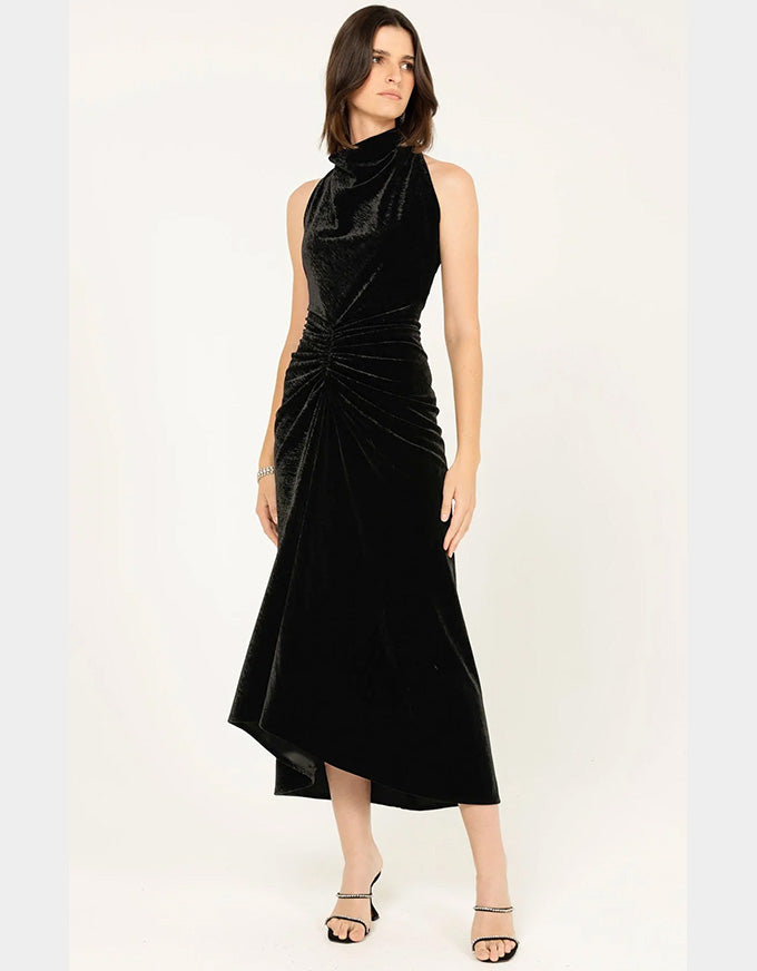A woman modeling a sleeveless black velvet midi dress with a high draped neckline, gathered waist, and asymmetrical hem, photographed against a plain light background.