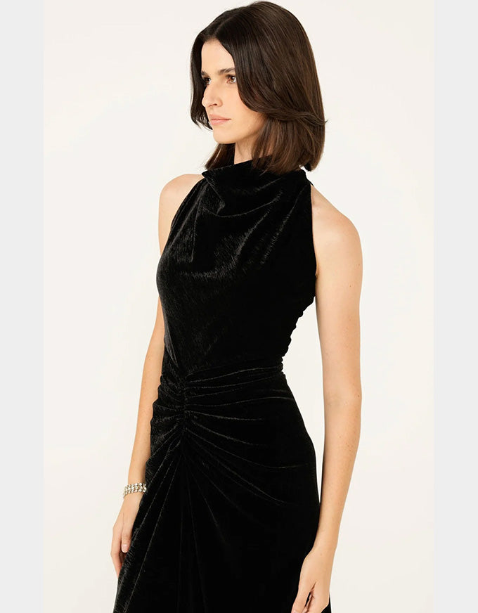 A woman wearing a sleeveless black velvet dress with a high mock neck and gathered ruched skirt, photographed against a plain light background.