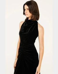 A woman wearing a sleeveless black velvet dress with a high mock neck and gathered ruched skirt, photographed against a plain light background.