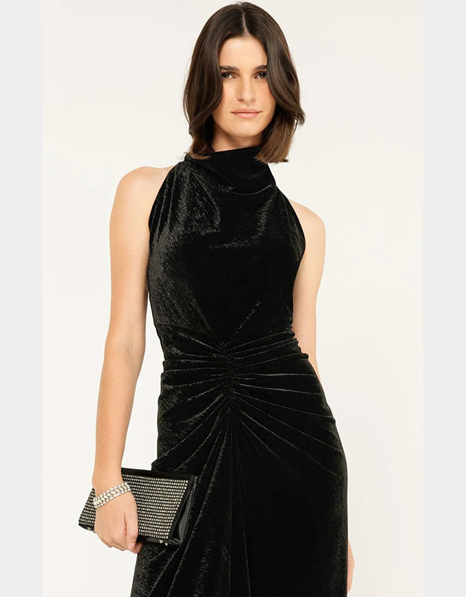 A woman modeling a sleeveless black velvet dress with a high mock neck and gathered ruching at the waist, holding a black studded clutch against a plain light background.