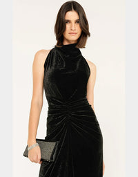 A woman modeling a sleeveless black velvet dress with a high mock neck and gathered ruching at the waist, holding a black studded clutch against a plain light background.