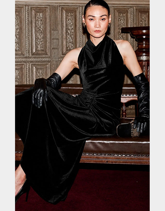 A model wearing a black velvet halter-neck dress with matching black leather gloves, seated on a wooden bench against an ornate wood-paneled wall.