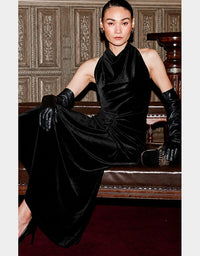 A model wearing a black velvet halter-neck dress with matching black leather gloves, seated on a wooden bench against an ornate wood-paneled wall.
