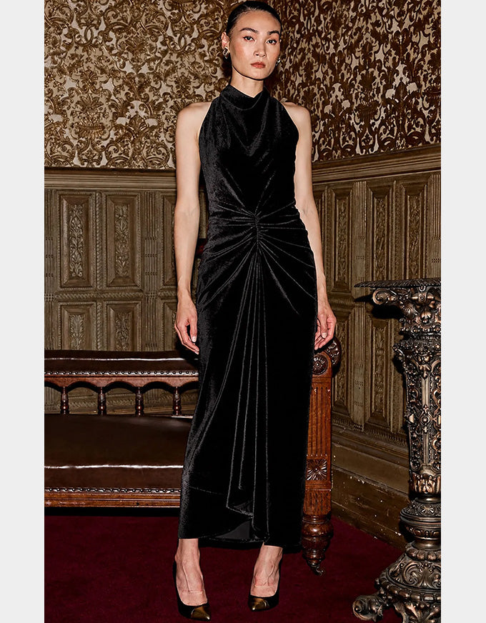 A model wearing a sleeveless black velvet high-neck maxi dress with a gathered ruched waist, standing in an ornate vintage room with patterned wallpaper and wood paneling.