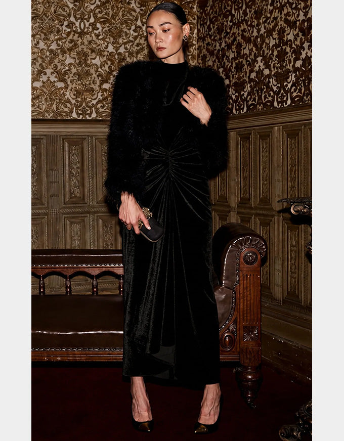 A woman stands in an ornate interior wearing a long black velvet dress with a gathered waist, a black fur-look jacket, and black pointed heels. She holds a small black clutch against a background of patterned wallpaper and carved wood paneling.
