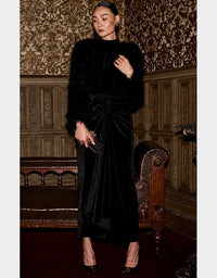 A woman stands in an ornate interior wearing a long black velvet dress with a gathered waist, a black fur-look jacket, and black pointed heels. She holds a small black clutch against a background of patterned wallpaper and carved wood paneling.