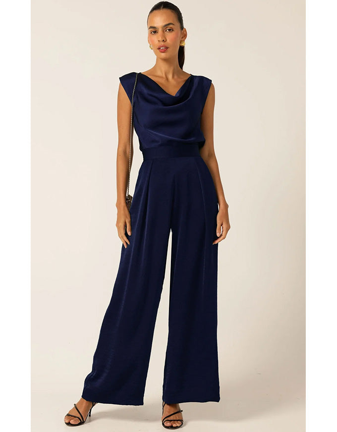 A woman modeling a navy blue sleeveless jumpsuit with a draped cowl neckline and wide legs, standing against a plain beige studio background.