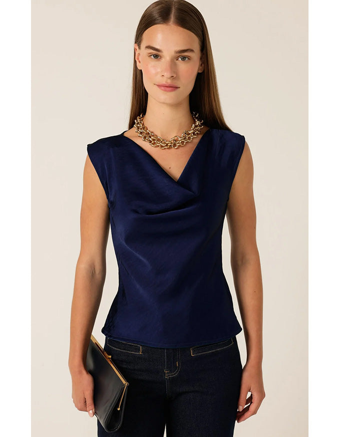 A woman modeling a sleeveless navy blue top with a draped V-neckline, paired with dark jeans and a chunky gold chain necklace, holding a black clutch against a light beige background.