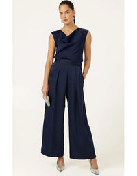 A woman modeling a navy blue sleeveless jumpsuit with a draped neckline, a matching waistband, and wide-leg cropped pants, paired with silver pointed-toe heels against a plain off-white studio background.