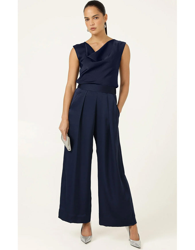 A woman modeling a navy blue sleeveless jumpsuit with a draped neckline, a matching waistband, and wide-leg cropped pants, paired with silver pointed-toe heels against a plain off-white studio background.