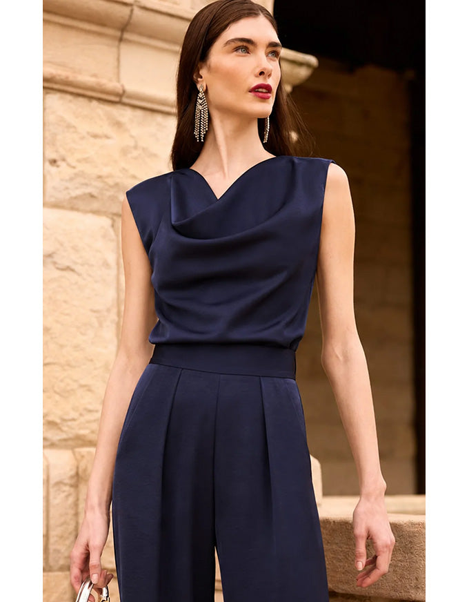 A woman wearing a navy blue sleeveless jumpsuit with a draped cowl neckline and wide legs, standing outdoors against a beige stone wall.