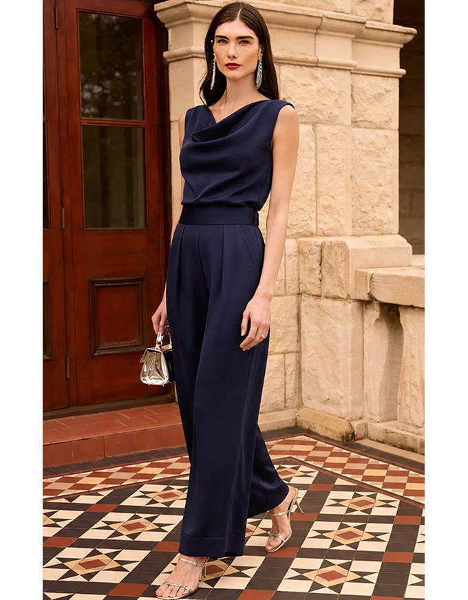 A woman models a navy blue sleeveless cowl-neck jumpsuit in an outdoor setting with a stone pillar and patterned tile floor.