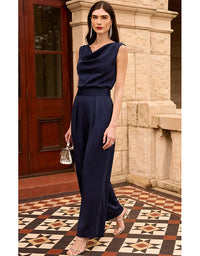 A woman models a navy blue sleeveless cowl-neck jumpsuit in an outdoor setting with a stone pillar and patterned tile floor.