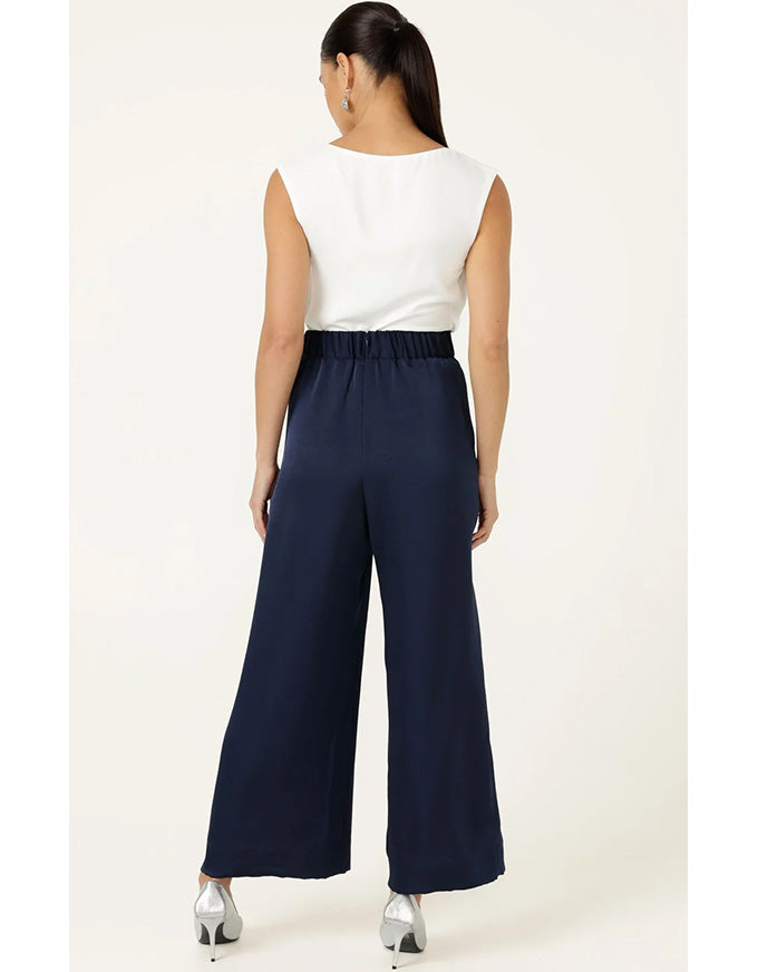 A woman shown from the back wearing navy blue wide-leg trousers and a white sleeveless top, standing against a plain light beige studio background.