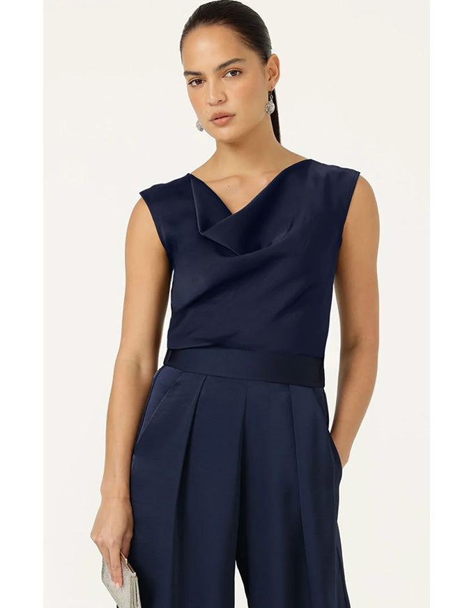 A woman modeling a navy blue sleeveless jumpsuit with a draped cowl neckline, a matching waistband, and wide-leg pleated trousers, set against a plain light beige background.