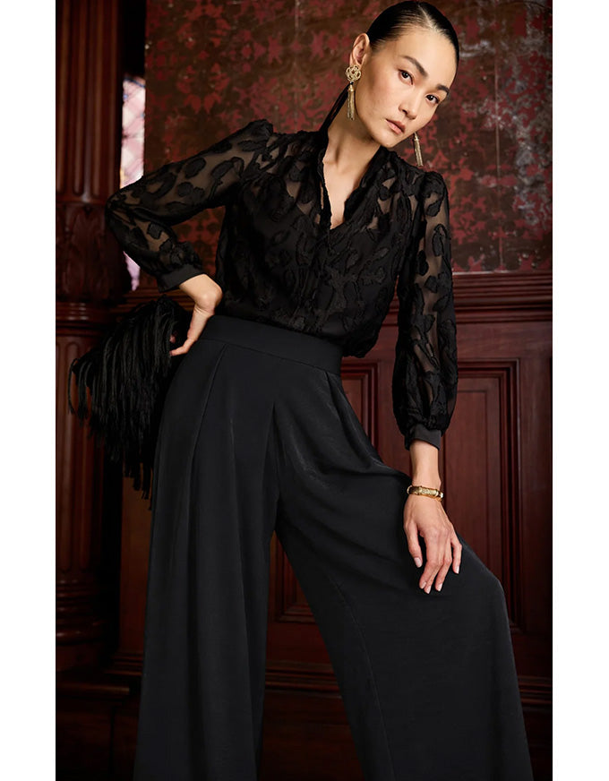 Woman wearing a black sheer blouse and wide-leg pants against a dark wooden paneled wall.