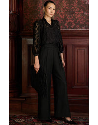 Woman in a black outfit with a fringed bag standing against a wooden paneled wall.
