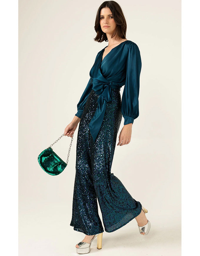 Woman wearing a teal outfit with sequin pants and a matching top, holding a green handbag on a white background.