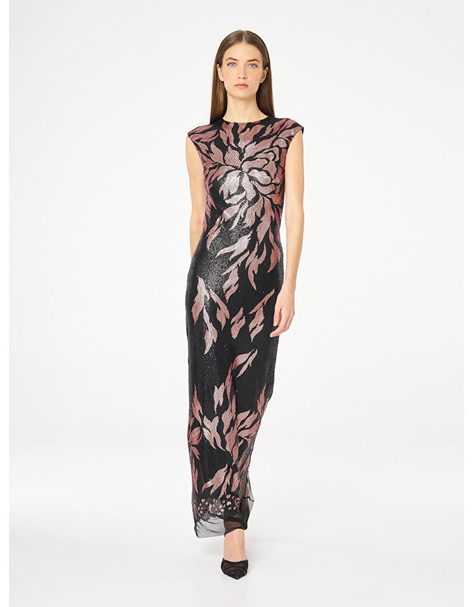A woman modeling a sleeveless, floor-length black dress with a large pink and gray floral pattern, photographed against a plain white studio background.