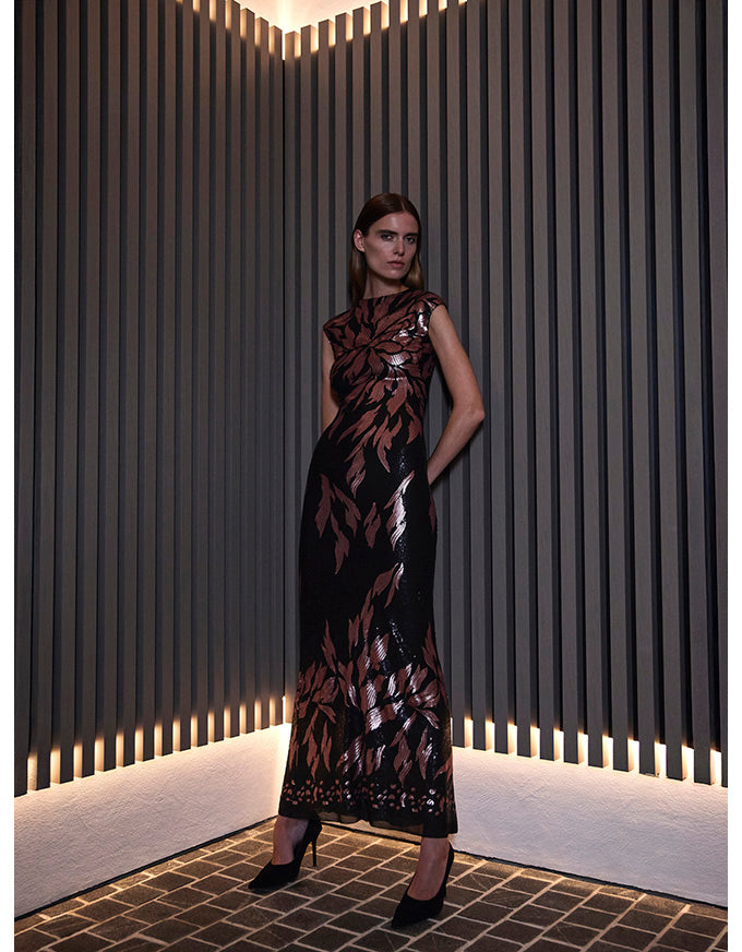 A woman models a sleeveless black midi dress with a metallic rose gold leaf pattern, standing in a modern room with vertical slatted walls.
