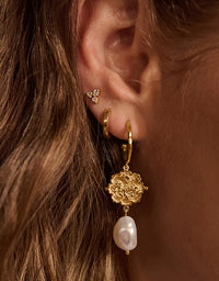 Close-up of an ear wearing gold hoop earrings with a pearl drop and floral design.