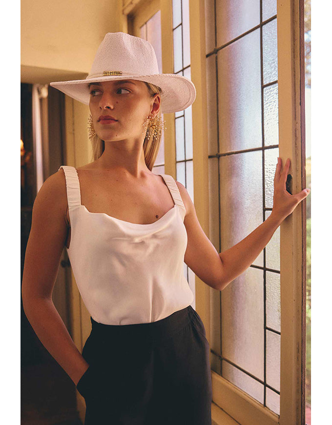 A woman wearing a white wide-brim hat and a white sleeveless top with black bottoms, standing indoors next to a window with a warm, golden-toned background.
