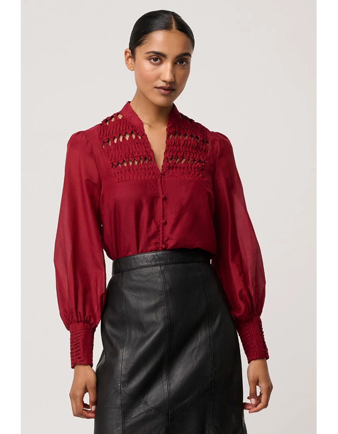 Woman wearing a red blouse with intricate detailing and black leather pants on a plain background