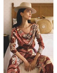 Woman in a patterned dress and hat sitting on a bed in a bedroom setting.