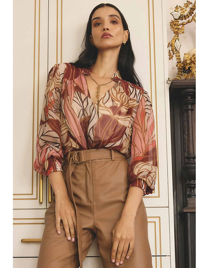 Woman wearing a patterned blouse and beige pants in an elegant interior setting.