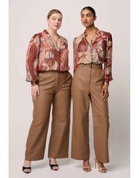 Two women wearing patterned tops and brown pants on a plain background