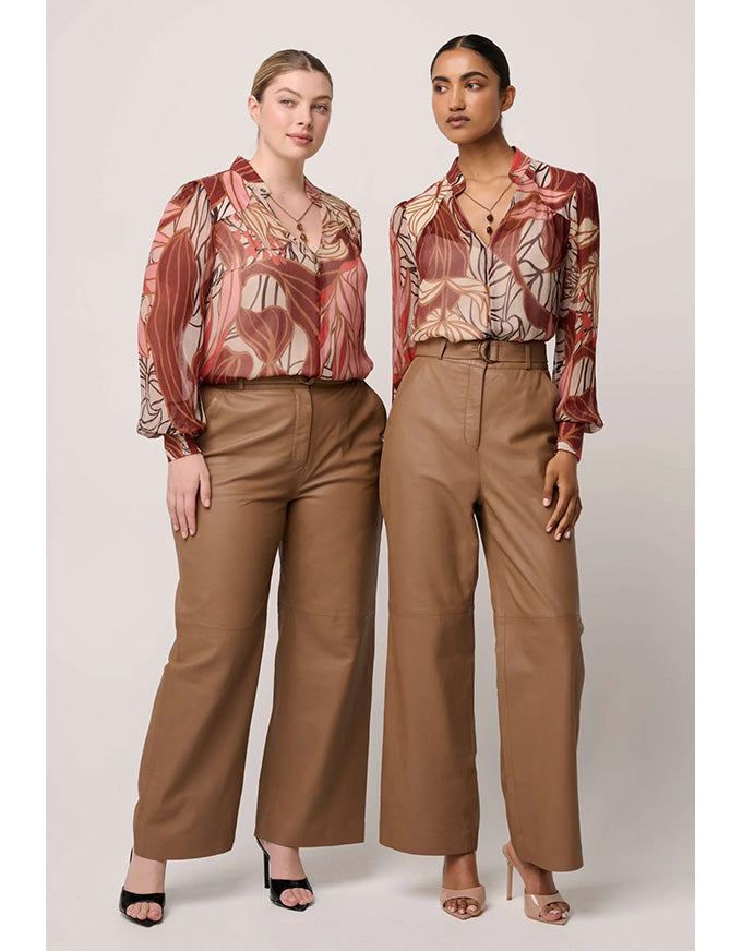 Two women wearing patterned tops and brown pants on a plain background