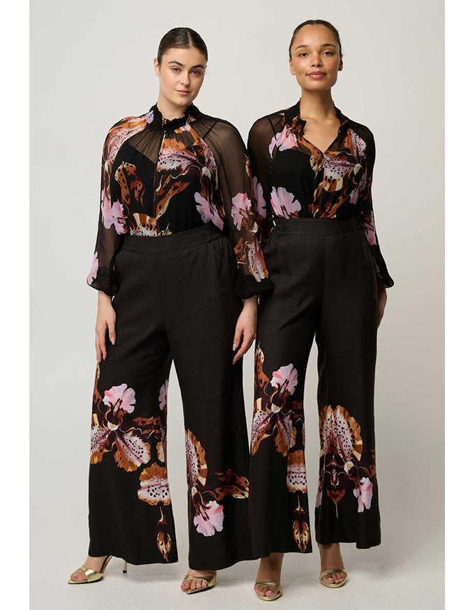 Two models wearing matching black wide-leg trousers and sheer long-sleeve tops with a brown, pink, and orange floral print, standing against a light gray studio background.