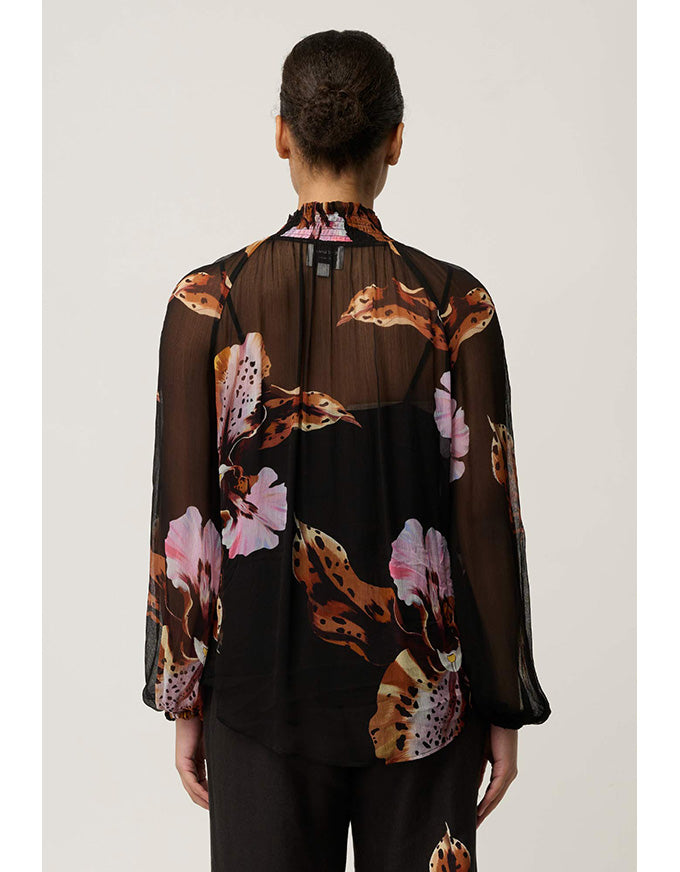 A model viewed from the back wearing a sheer black long-sleeve blouse with a brown, pink, and beige floral print, paired with black trousers, against a light gray studio background.
