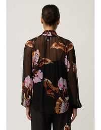 A model viewed from the back wearing a sheer black long-sleeve blouse with a brown, pink, and beige floral print, paired with black trousers, against a light gray studio background.