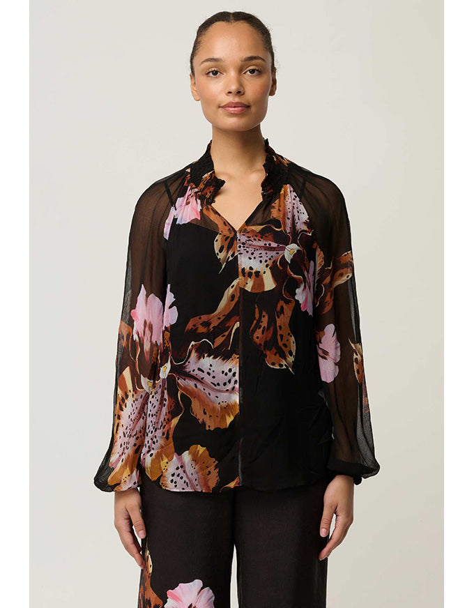 A woman modeling a sheer black long-sleeve blouse with a bold brown, pink, and beige floral and animal-inspired print, paired with black trousers, against a light gray studio background.