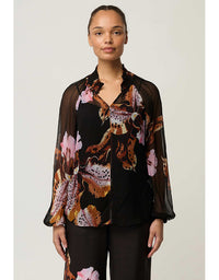 A woman modeling a sheer black long-sleeve blouse with a bold brown, pink, and beige floral and animal-inspired print, paired with black trousers, against a light gray studio background.