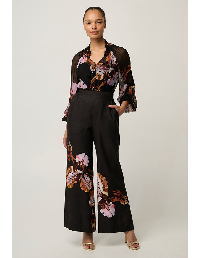 A woman modeling a black wide-leg jumpsuit with a sheer long-sleeve top and pink, purple, and brown floral print, paired with beige heeled sandals, against a light gray studio background.