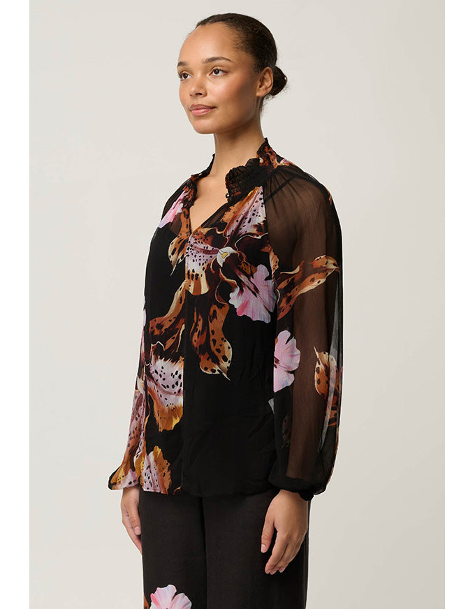 A woman modeling a black long-sleeve blouse with sheer mesh sleeves and a bold brown, pink, and white floral print, paired with black pants against a light gray studio background.