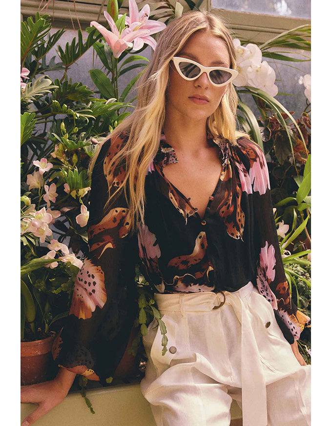 A woman wearing a black long-sleeve blouse with a colorful bird and floral print, paired with white trousers and white cat-eye sunglasses, posed in front of a lush background of green plants and pink and white flowers.