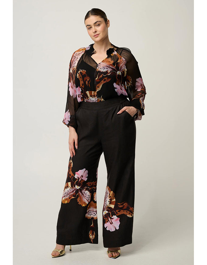 A woman models a black wide-leg pants set with a matching sheer long-sleeve top featuring a brown, pink, and orange floral print, photographed against a neutral light gray studio background.