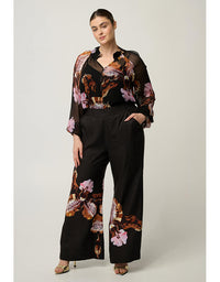 A woman models a black wide-leg pants set with a matching sheer long-sleeve top featuring a brown, pink, and orange floral print, photographed against a neutral light gray studio background.