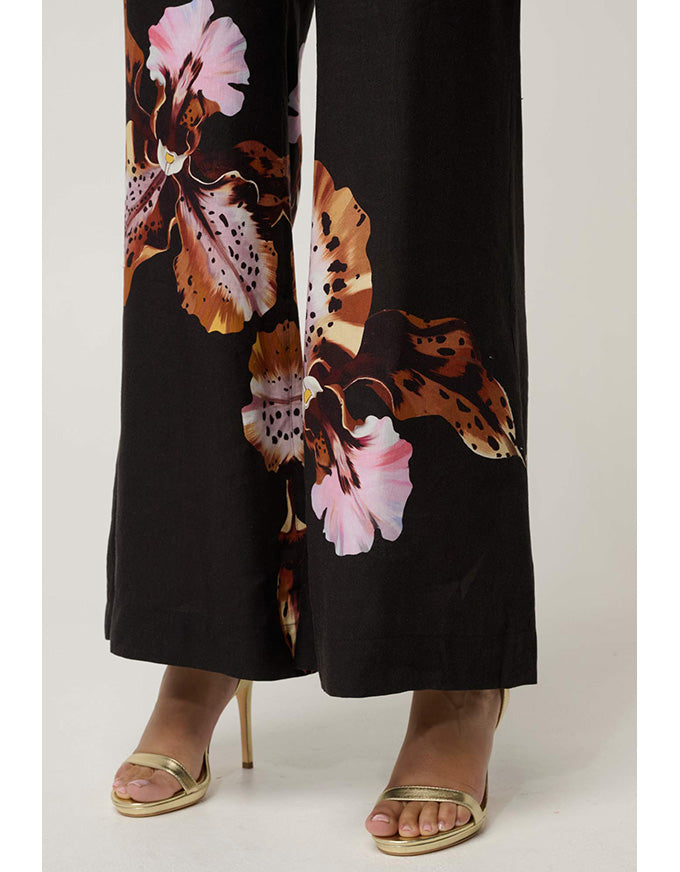 Black wide-leg pants with a large brown, orange, and pink floral print, shown on a model wearing gold heeled sandals against a light gray studio background.