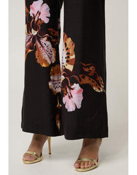 Black wide-leg pants with a large brown, orange, and pink floral print, shown on a model wearing gold heeled sandals against a light gray studio background.