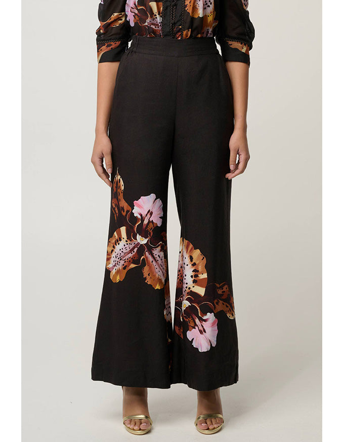 A model wearing black wide-leg pants with a large brown, orange, and pink floral print on the lower legs, paired with a matching patterned top and beige sandals, against a light gray studio background.